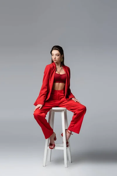 Full length of brunette young woman in red suit and shoes sitting on white chair on dark grey — Stock Photo
