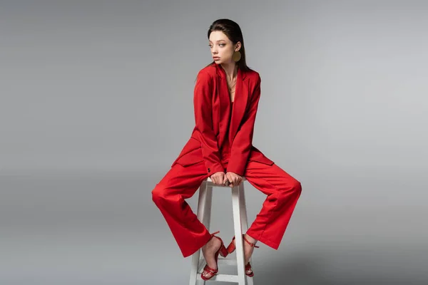 Full length of brunette woman in red suit and earring sitting on white chair on dark grey — Stock Photo