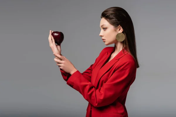 Vista lateral del joven modelo en traje rojo posando con manzana aislada sobre gris - foto de stock