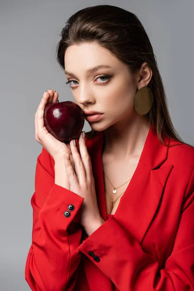 Mujer joven en blazer rojo y pendiente con manzana aislada en gris - foto de stock