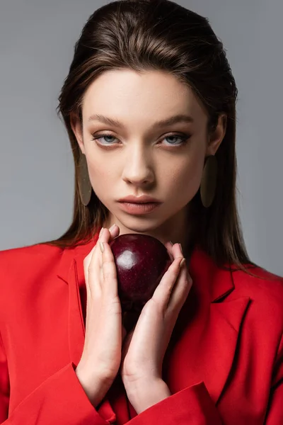 Молода жінка в червоному блістері і сережки тримає яблуко ізольовано на сірому — стокове фото