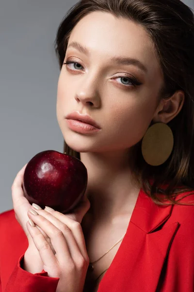 Pretty woman in red blazer and earring holding ripe apple isolated on grey — Stock Photo