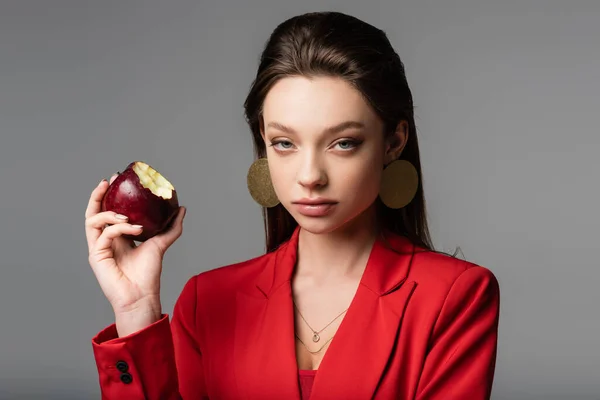 Mujer joven de moda en blazer rojo y pendientes con manzana aislada en gris - foto de stock