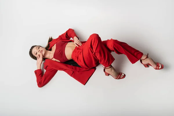 Vista de ángulo alto de la mujer joven con estilo en traje rojo y zapatos acostados en gris - foto de stock