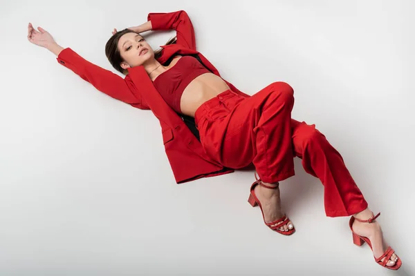 Vista de ángulo alto de elegante modelo joven en traje rojo y zapatos acostados en gris - foto de stock