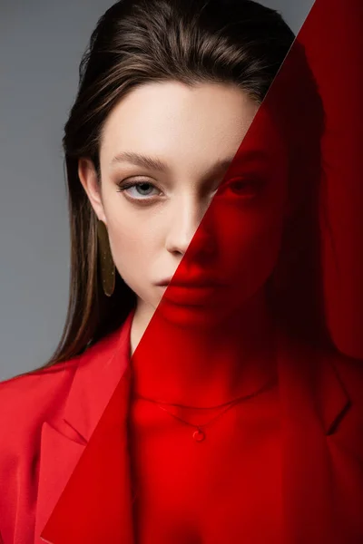 Mujer joven de moda mirando a la cámara a través de vidrio rojo aislado en gris - foto de stock