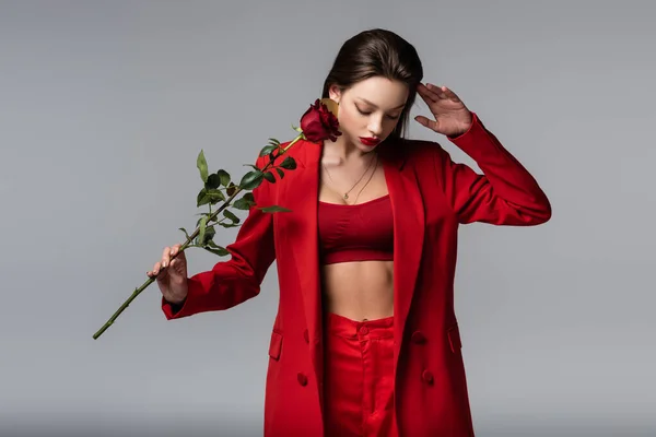 Modelo joven en traje rojo posando con rosa aislada sobre gris - foto de stock