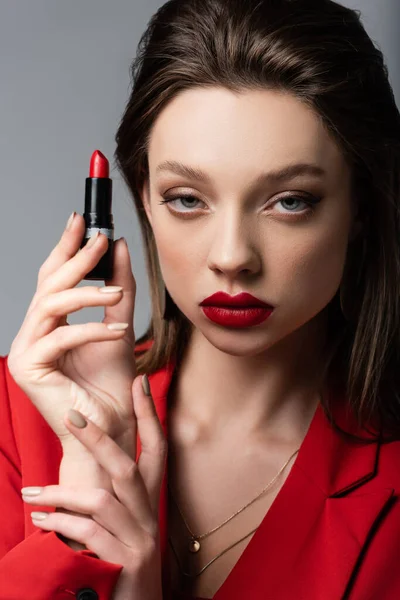 Primer plano de la joven en blazer rojo sosteniendo lápiz labial aislado en gris - foto de stock