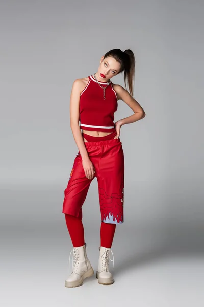 Longueur totale de la jeune femme en tenue de sport rouge et bottes posant avec la main sur la hanche sur gris — Photo de stock