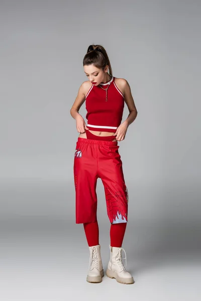 Longitud completa de modelo joven en traje deportivo rojo y botas posando en gris - foto de stock