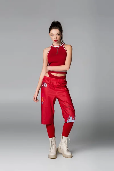 Pleine longueur de jolie femme en tenue de sport rouge et bottes posant sur gris — Photo de stock