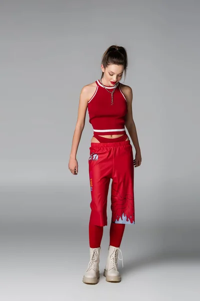 In voller Länge stylische junge Frau in rotem sportlichem Outfit und Stiefeln posiert auf grau — Stockfoto