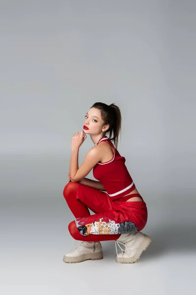 In voller Länge stilvolle junge Frau in rotem, sportlichem Outfit und Stiefeln sitzend, während sie auf grau posiert — Stockfoto