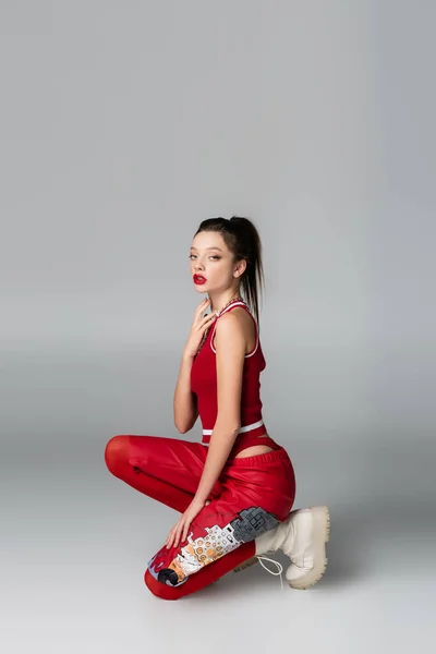 Full length of trendy young woman in red sportive outfit and boots sitting while posing on grey — Stock Photo