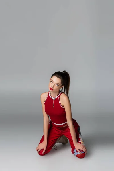 Модная молодая женщина в красном спортивном костюме, стоящая на коленях на сером — стоковое фото