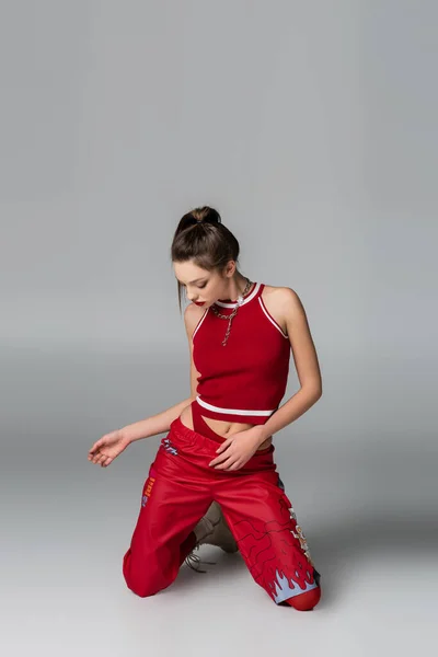 Стильная молодая женщина в красном спортивном костюме, стоящая на коленях на сером — стоковое фото