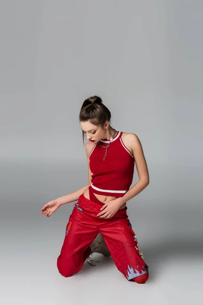 Mujer joven con estilo en traje deportivo rojo de pie sobre las rodillas en gris - foto de stock