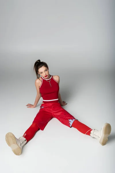 Longueur totale de la jeune femme en tenue de sport rouge et des bottes assises sur gris — Photo de stock