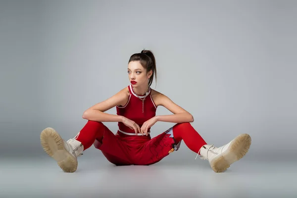 Full length of trendy young model in red sportive outfit and boots sitting and posing on grey — Stock Photo