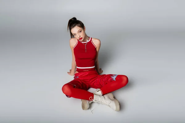 Jeune modèle tendance en tenue de sport rouge et bottes assises avec jambes croisées sur gris — Photo de stock