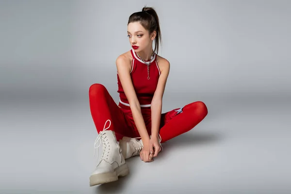 Junges stylisches Model in rotem sportlichem Outfit und Stiefeln auf grau sitzend — Stockfoto