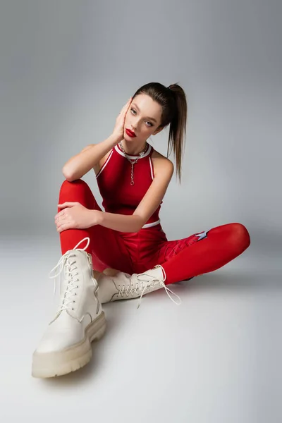 Mujer joven y elegante en traje deportivo rojo y botas sentadas en gris - foto de stock