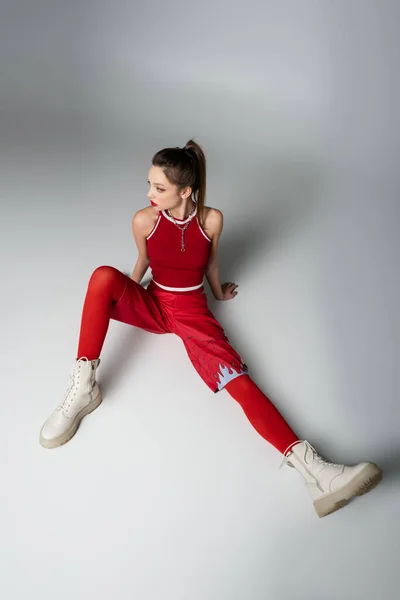 Высокий угол обзора молодой стильной женщины в красном спортивном костюме и ботинках, сидящих на сером — стоковое фото