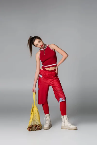 Piena lunghezza di giovane donna in abito sportivo rosso tenuta borsa stringa con le mele mentre posa con mano sul fianco sul grigio — Foto stock