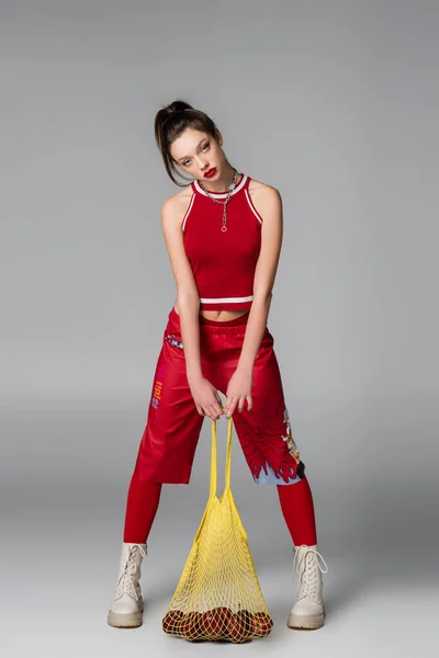 Longitud completa de la mujer joven de moda en traje deportivo rojo sosteniendo bolsa de cadena con manzanas en gris - foto de stock