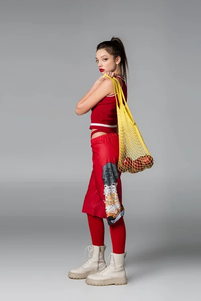 Full length of trendy young model in red sportive outfit holding string bag with apples on grey — Stock Photo