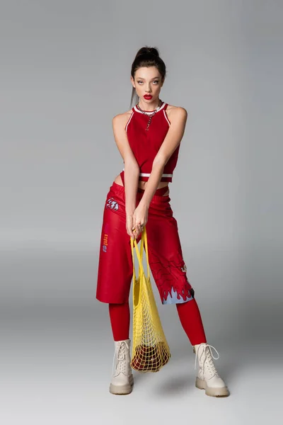 Longitud completa del modelo de moda en traje deportivo rojo que sostiene el bolso de cuerda con manzanas en gris - foto de stock