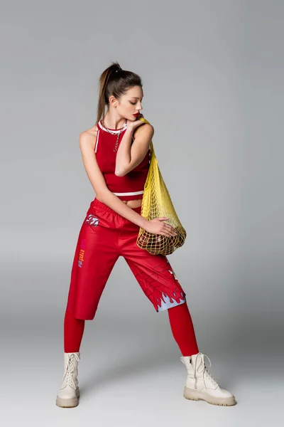 Full length of trendy woman in red sportive outfit holding yellow string bag with apples on grey — Stock Photo