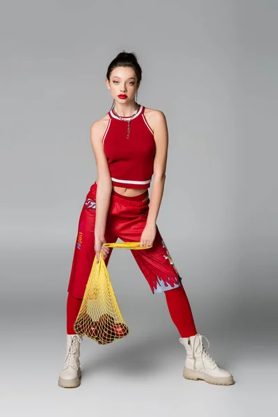 Full length of young trendy woman in red sportive outfit holding yellow string bag with ripe apples on grey — Stock Photo