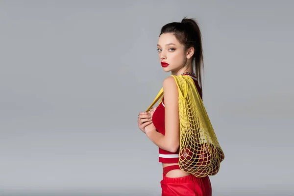 Young stylish woman in red sportive outfit holding string bag with apples isolated on grey — Stock Photo