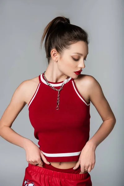 Mujer joven y elegante en traje deportivo rojo y cadena de collar aislado en gris - foto de stock