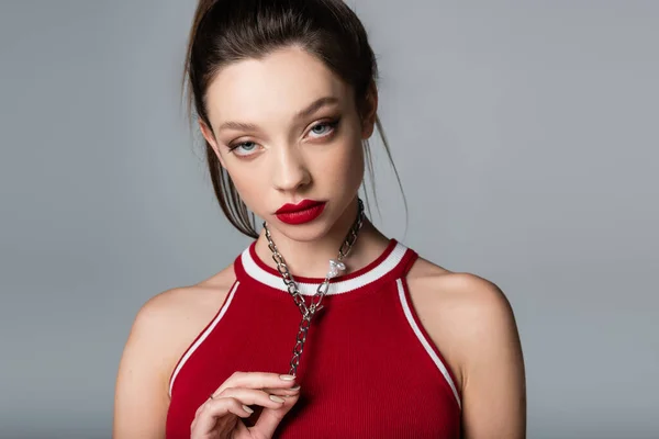 Молода стильна жінка з червоними губами торкається намиста ланцюжка ізольовані на сірому — стокове фото