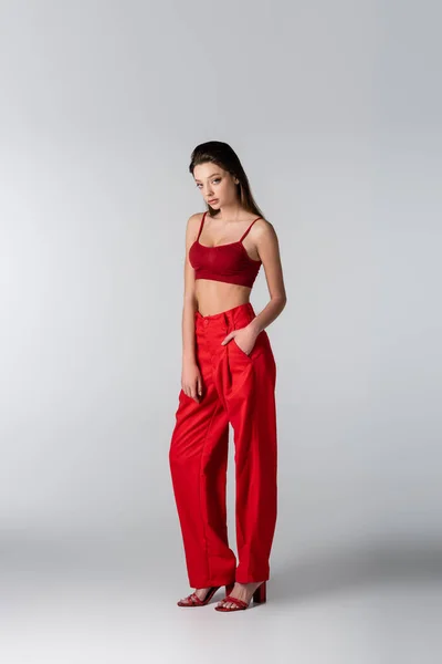 Longitud completa de la mujer joven en top rojo de la cosecha y pantalones posando con la mano en el bolsillo en gris - foto de stock