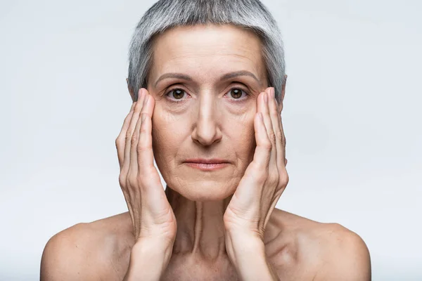 Mujer de mediana edad con el pelo gris tocando la cara con arrugas aisladas en gris - foto de stock