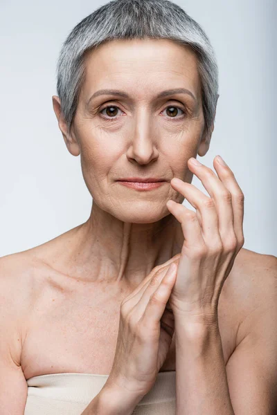 Mujer de mediana edad con hombros desnudos tocando las manos aisladas en gris - foto de stock