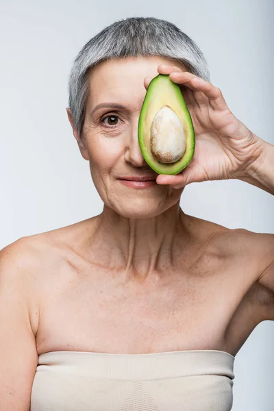 Женщина средних лет покрывает лицо половинкой авокадо, изолированной на сером — стоковое фото