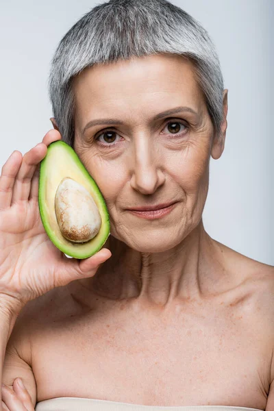 Женщина средних лет, держащая половину свежего авокадо, изолированного на сером — стоковое фото