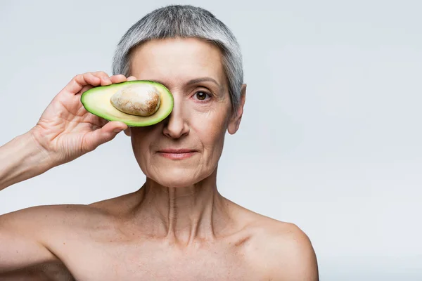 Femme d'âge moyen couvrant l'œil avec la moitié de l'avocat isolé sur gris — Photo de stock