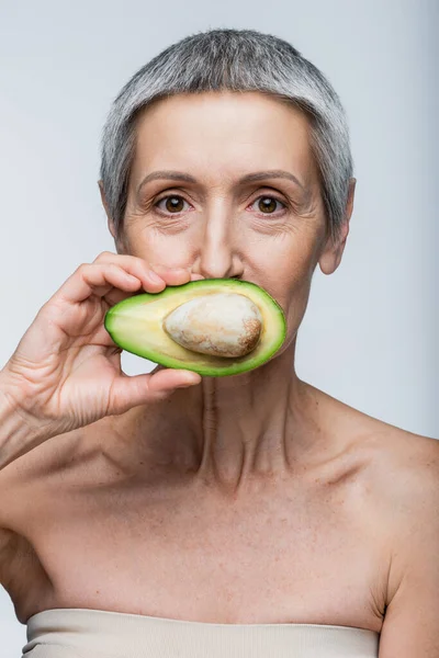 Женщина средних лет покрывает рот половинкой авокадо, изолированной на сером — стоковое фото