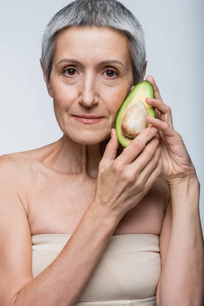 Женщина средних лет, держащая половину органического авокадо, изолированного на сером — стоковое фото