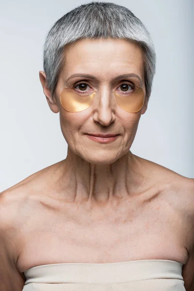 Middle aged woman in golden eye patches looking at camera isolated on grey — Stock Photo