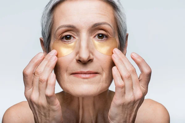 Middle aged woman applying golden eye patches while looking at camera isolated on grey — Stock Photo