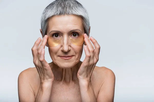 Middle aged woman applying golden eye patches and looking at camera isolated on grey — Stock Photo