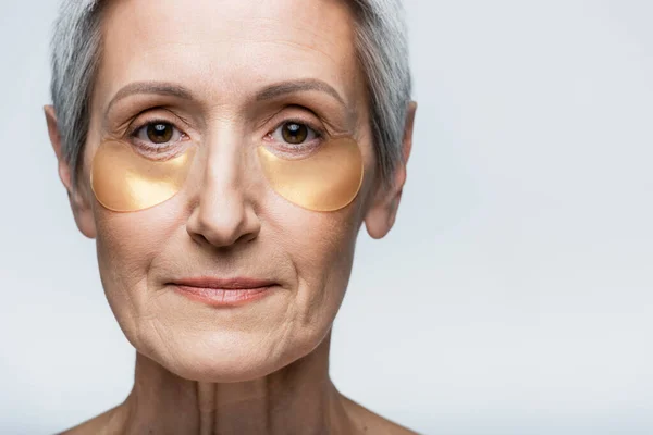 Close up of middle aged woman in collagen eye patches looking at camera isolated on grey — Stock Photo