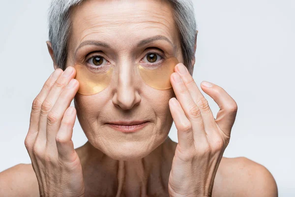 Mulher de meia idade aplicando manchas de hidrogel nos olhos e olhando para a câmera isolada em cinza — Fotografia de Stock