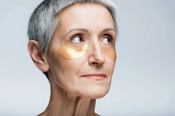Middle aged woman in golden eye patches looking away isolated on grey — Stock Photo